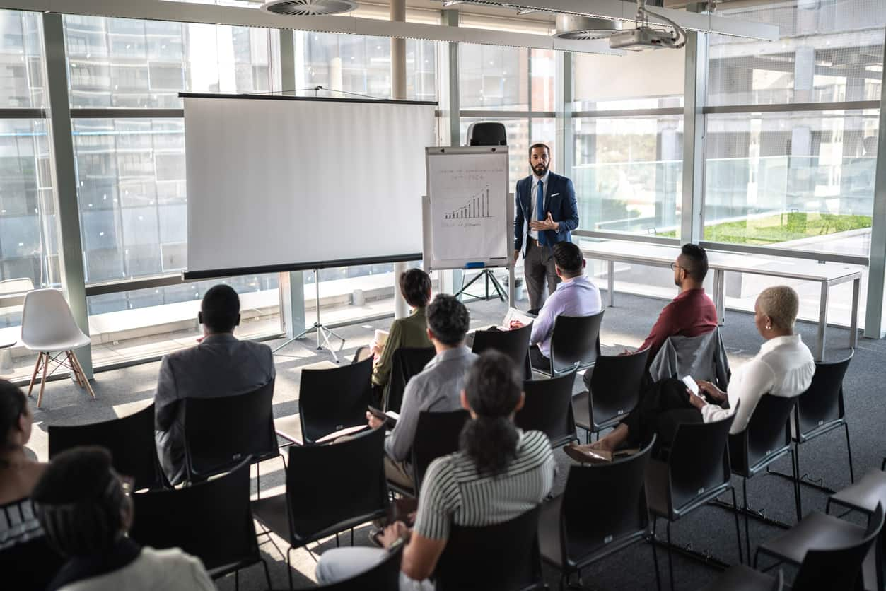 Man standing with a board talking to a group of seated people 