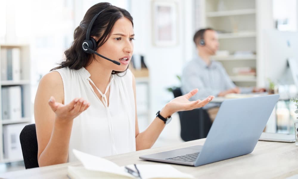 A woman wearing a headset shrugging at her laptop in frustration.