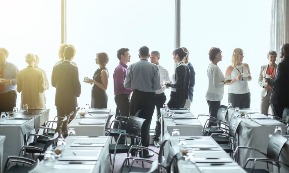 A group of people standing by the windows of a conference room and socializing.